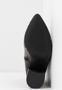 Suola di una scarpa nera a punta con motivo testurizzato, appoggiata verticalmente contro un angolo di muro e superficie bianchi.