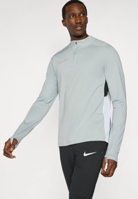 Ανοιχτό γκρι αθλητικό μακρυμάνικο μπλουζάκι με φερμουάρ στον γιακά, με χρυσό λογότυπο Nike και μαύρες πλαϊνές λεπτομέρειες, φορεμένο πάνω από μαύρο παντελόνι.