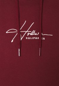 Sudadera burdeos con una textura suave, que presenta un logotipo bordado en blanco "Hollister California" y un cordón con puntas de metal.