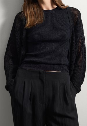 Femme portant un haut sans manches noir côtelé sous un cardigan noir en maille transparente avec un pantalon large noir taille haute, mains dans les poches.
