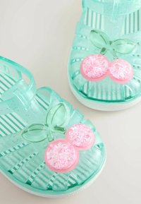 Sandales enfant translucides turquoise avec paillettes, ornées de décorations en forme de cerises roses avec des feuilles vertes sur les orteils.