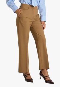 Pantaloni sartoriali color cammello con una texture liscia, design a gamba dritta, tasche laterali e vita media, abbinati a tacchi marroni con cinturini.