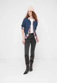 Γυναίκα που φοράει ροζ σκουφάκι, μπλε ζακέτα πάνω από ροζ πουκάμισο με καρό, λευκό τοπ, μαύρο στενό τζιν και ψηλά μαύρα μποτάκια με τακούνι, στέκεται μπροστά σε λευκό τοίχο.