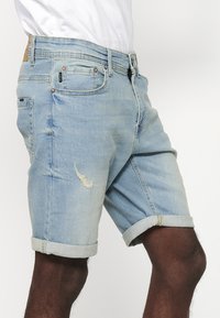 Shorts en denim bleu clair avec des ourlets roulés, présentant une texture usée et une légère décoloration. Comprend des poches avant et arrière.