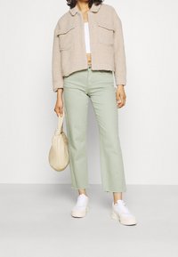 Beżowa kurtka o teksturowanym wykończeniu z dwoma przednimi kieszeniami, w połączeniu z jasnoszarymi, krótkimi dżinsami. Stylizacja dopełniają białe trampki i jasna torba.