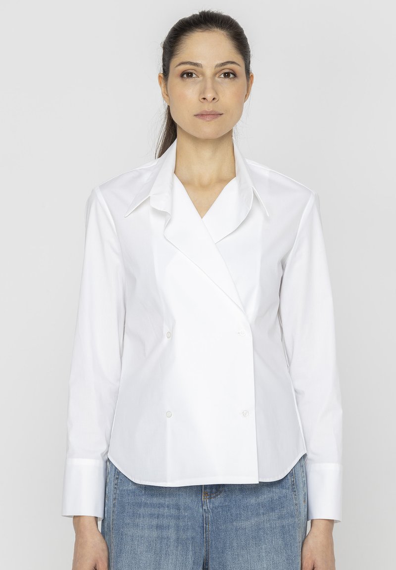 Biała, dwurzędowa koszula z wyłódkami, długimi rękawami i guzikami, w połączeniu z niebieskimi dżinsami. Gładka tekstura materiału.