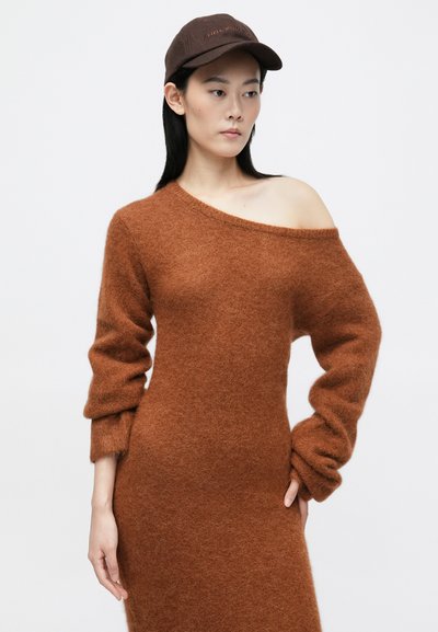 Rjavkasto oblačilo brez naramnic iz puhaste tkanine, z dolgimi, obsežnimi rokavi in prilegajočo silhueto. V kombinaciji s rjavim klobukom.