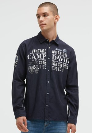 Junger Mann mit kurzen Haaren, der ein dunkelblaues Hemd mit Knöpfen und weißem Text mit Flugthema sowie Jeans trägt und nach links schaut.