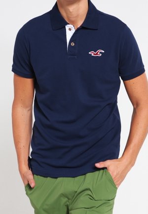 Granatowa koszulka polo z białym kołnierzykiem, z małym czerwonym logo na lewym ramieniu. Wykonana z miękkiej bawełny, z krótkimi rękawami.