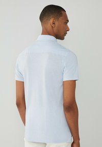 Svetlomodrá polokošeľa s krátkym rukávom vyrobená z mäkkého materiálu, so klasickým golierom a čistým dizajnom na zadnej strane bez viditeľného loga.