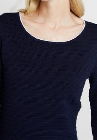 Haut bleu marine à manches longues avec un motif texturé, doté d'un col rond avec un bord contrastant blanc. Design simple et ajusté.