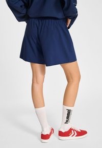 Shorts blu navy fino al ginocchio con elastico in vita, abbinati a scarpe da ginnastica rosse e calze bianche con scritte nere.