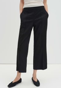 Pantalon noir sur mesure à jambe droite, doté de poches latérales et d'une texture lisse. Porté avec des chaussures plates noires, sur un fond neutre.