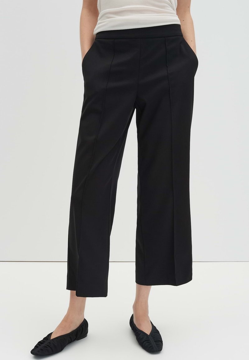 Pantalon noir sur mesure à jambe droite, doté de poches latérales et d'une texture lisse. Porté avec des chaussures plates noires, sur un fond neutre.
