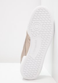 Vit gummisula på en beige Reebok-sneaker med diamantmönster och cirkulärt greppdetalj, placerad vertikalt mot en vit yta.