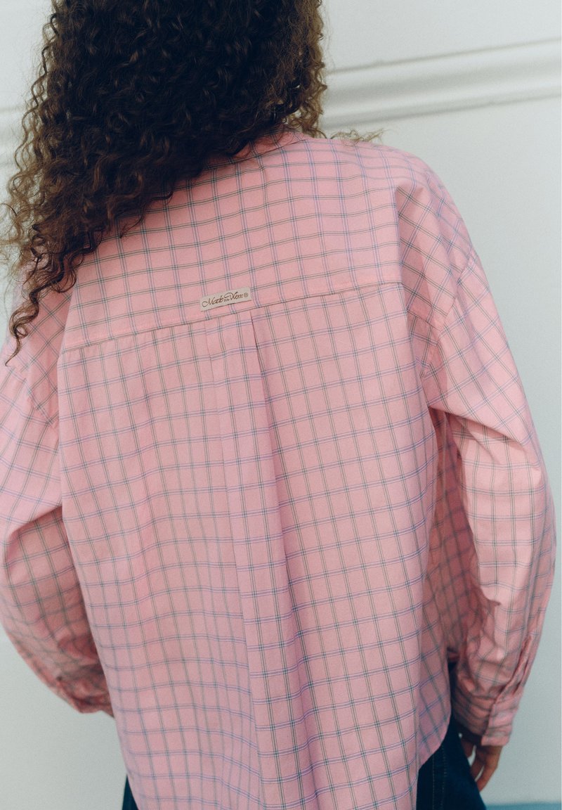 Personne aux longs cheveux bouclés portant une chemise ample à carreaux roses avec une petite étiquette rectangulaire dans le haut du dos.