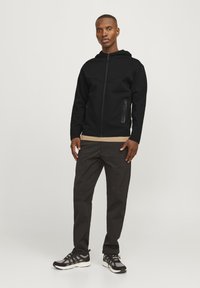 Svart zip-up hettegenser med en frontlomme og en glatt tekstur, paret med mørke, tapered bukser og svarte sneakers med grå detaljer.