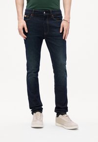 Slim fit σκοτεινές μπλε τζιν παντελόνες φορεμένες με ανοιχτό μπεζ αθλητικά παπούτσια και πράσινη μπλούζα, με τα πόδια και τα χέρια να είναι χαλαρωμένα στο πλάι.