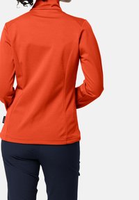 Kobieta w dopasowanej pomarańczowej kurtce z długim rękawem i ciemnych spodniach, widziana z tyłu, z kręconymi włosami związanymi w kok.