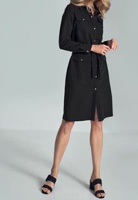 Schwarzes Hemdkleid mit langen Ärmeln, zwei Brusttaschen, einem Taillengürtel und einem Schlitz vorne. Kombiniert mit schwarzen Sandalen.