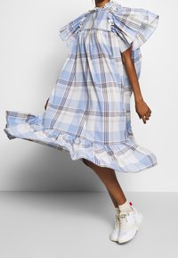 Žena oblečená v splývavých šatech v modro-bílé kostkované vzoru s volantovými rukávy a lemem, doplněná bílými teniskami a béžovými ponožkami.