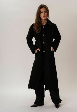 Hosszú barna hajú nő, fekete hosszú kabátot visel fekete nadrág és hegyes fekete cipők fölött, kezét a kabátzsebekben tartva áll.