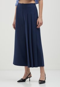 Pantaloni a gamba larga plissettati di colore navy, realizzati in tessuto morbido, con vita alta e zip laterale nascosta. Indossati con décolleté nere a punta.