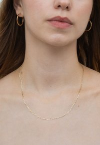 Halskette mit goldfarbener Kette und verlängerten Gliedern, um den Hals getragen, kombiniert mit passenden kleinen Creolen. Glatte Textur.