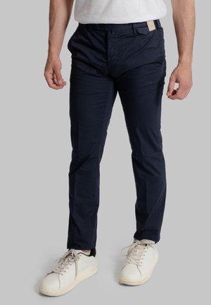 Uomo che indossa pantaloni blu navy aderenti e scarpe da ginnastica bianche, in piedi davanti a uno sfondo grigio chiaro uniforme.