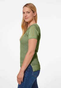 Grünes, kurzärmliges Strickoberteil mit strukturiertem Design und dekorativen Nähten an den Schultern, kombiniert mit blauen Jeans.
