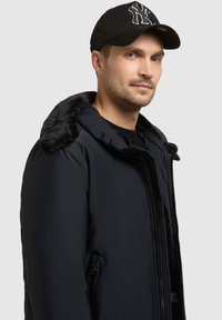 Mężczyzna w czarnej zimowej kurtce z futrzanym kołnierzem i czapce baseballowej w kolorze czarnym z białym emblematem, lekko patrzy w prawo na jasnoszarym tle.