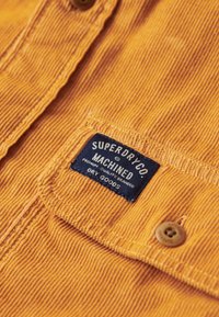 Kortærmet orange kordfløjlsskjorte med synlige stikninger, en brun knap og et blåt mærke med teksten "SUPERDRY CO. MACHINED DRY GOODS."