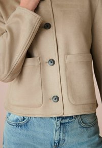 Veste en daim beige avec un col, quatre fermetures à boutons noirs et deux poches avant. Associée à un jean en denim bleu clair.