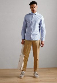  Lichtblauwe overhemdd, kaki broek en grijze schoenen. Houdt een witte tote bag vast. Glad textuur en eenvoudig ontwerp.