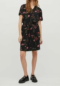 Sort blomsterkjole med korte ærmer, V-hals, indsnævret talje med bælte og lyserøde og grønne blomsterprint. Kombineret med sorte blanke sko.