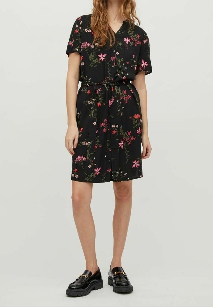 Sort blomsterkjole med korte ærmer, V-hals, indsnævret talje med bælte og lyserøde og grønne blomsterprint. Kombineret med sorte blanke sko.