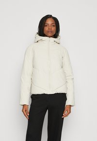 Polstret beige jakke med hette, som har glidelås foran, elastisk kant og chevronsøm. Brukt over svarte bukser.