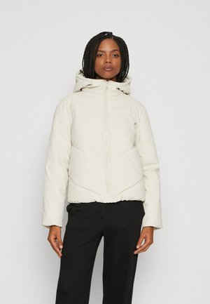 Veste beige rembourrée avec capuche, dotée d'une fermeture éclair à l'avant, d'un ourlet élastiqué et de surpiqûres en forme de chevrons. Portée sur un pantalon noir.