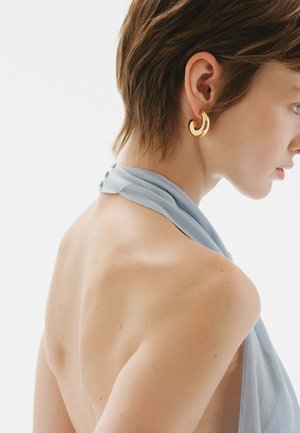 Mujer joven con cabello corto que lleva un top halter azul claro y grandes pendientes de aro dorados, de perfil contra un fondo liso.