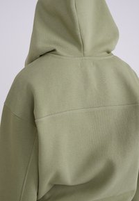 Felpa verde oliva con cappuccio a marsupio, realizzata in un morbido tessuto misto cotone, con una vestibilità comoda, cappuccio regolabile e dettagli di cucitura visibili.