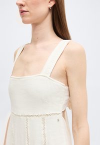 Γυναίκα με αμάνικο μπεζ φόρεμα με φαρδιές τιράντες και διακοσμητικές ραφές, απεικονιζόμενη από τους ώμους μέχρι το μέσο του κορμού.