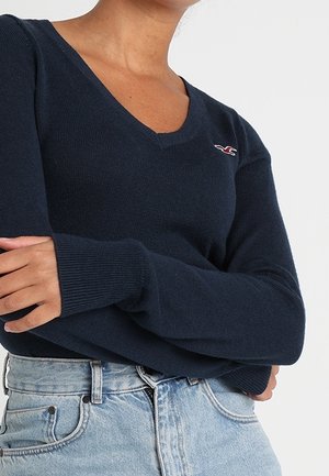 Námořnická modrá svetr s výstřihem do V z měkkého pleteného materiálu, s dlouhými rukávy a malým logem na hrudi. Vytvořeno v kombinaci s džíny z jasného denimového materiálu s vysokým pasem.