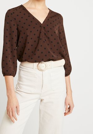 Kobieta ubrana w brązową bluzkę z długim rękawem w czarne kropki, włożoną do białych spodni z wysokim stanem i białym paskiem z okrągłą klamrą.