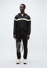 Musta vetoketjullinen takki, jossa valkoiset ja neonvihreät raidat, yhdistetty mustiin housuihin ja valkoisiin lenkkareihin. Pitää kädessään tennismailaa.