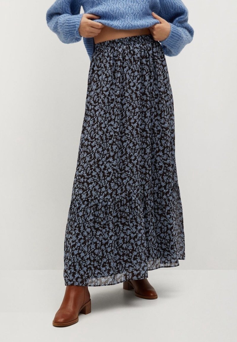 Personne portant une longue jupe noire avec un petit imprimé floral bleu, un pull bleu et des bottines marron, debout devant un fond blanc uni.