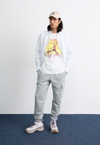 White and gray striped shirt over a graphic t-shirt, gray sweatpants, and light sneakers with red accents, complemented by a beige cap.