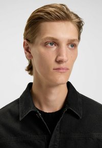 Zwarte denimjack met een kraag, met zichtbare stiksels en een gestructureerde stof. Draagt over een zwarte trui, tegen een neutrale achtergrond.