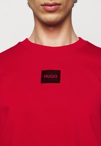 Homme portant une chemise rouge vif avec un patch noir marqué "HUGO" centré sous l'encolure, cadré au niveau du menton et des épaules.
