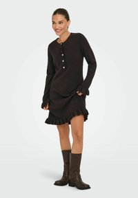 Kvinna som ler, iklädd en mörkbrun långärmad klänning med rufsiga fållar och manschetter, tillsammans med knähöga bruna skinnstövlar.
