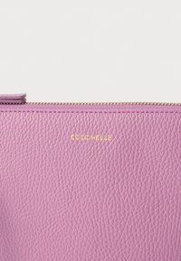 Lila, texturált bőrből készült erszény arany cipzárral és a "Coccinelle" márkanév domborítva az elején. Simára kidolgozott felület enyhe fényes csillogással.